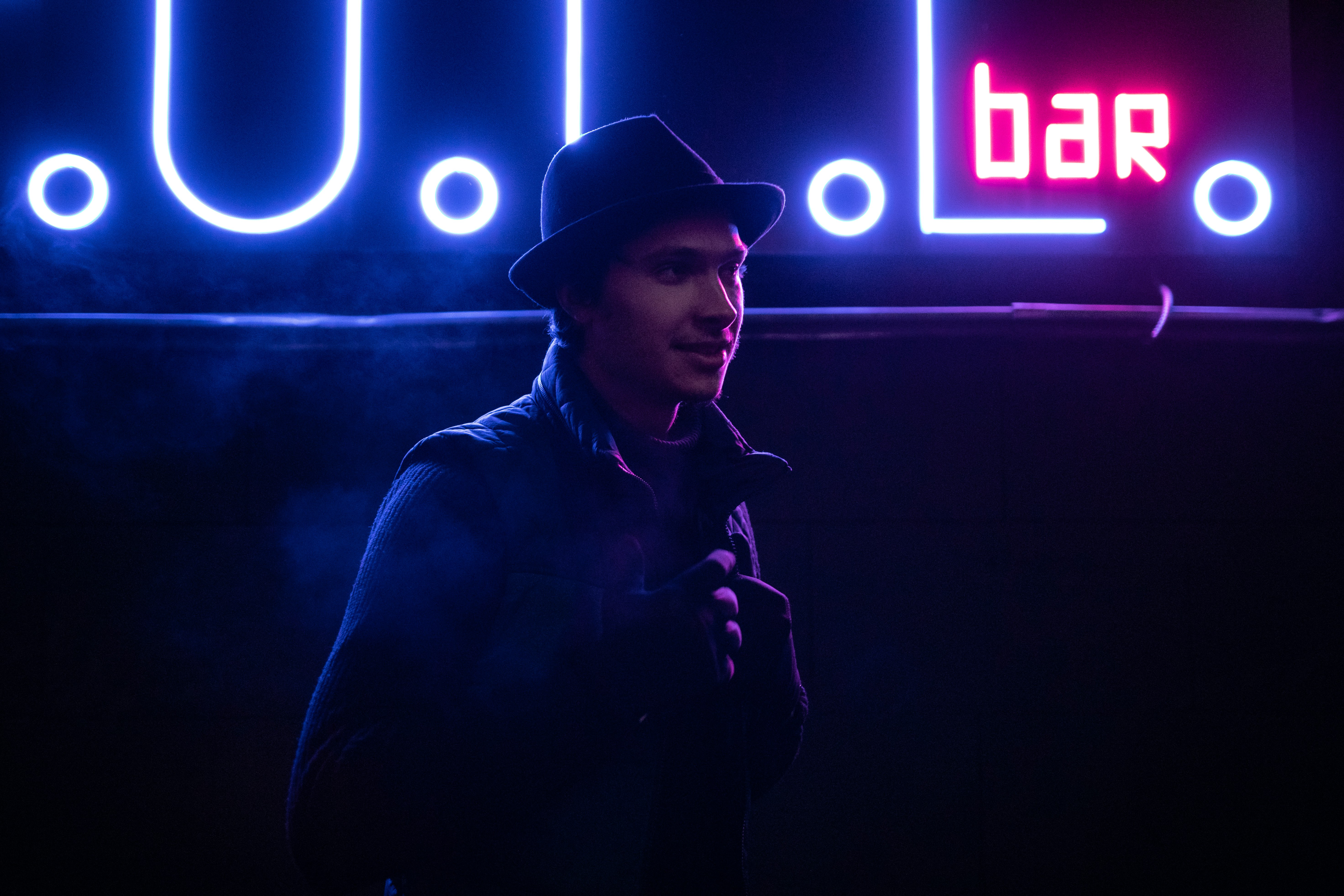 man in black leather jacket wearing black hat standing near blue and red open neon signage